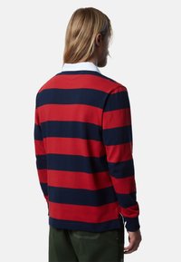 Man met lang haar in een rood en marineblauwe gestreepte long-sleeve shirt en groene broek, bekeken van achteren tegen een effen achtergrond.