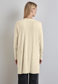 Długi, jasnobeżowy kardigan z prążkowanymi rękawami, o luźnym kroju i zaokrąglonym hemem, wykonany z miękkiego materiału.