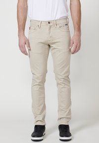 Jeans beige de corte slim, elaborados con una mezcla de algodón, que presentan una cremallera con cierre de botón, cinco bolsillos y detalles de costura sutiles.