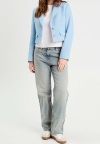 Hellblaue, strukturierte, kurz geschnittene Jacke mit zwei silbernen Knöpfen, kombiniert mit einem weißen T-Shirt und hellblauen, zeitlos geschnittenen Jeans sowie weißen Sneakers.