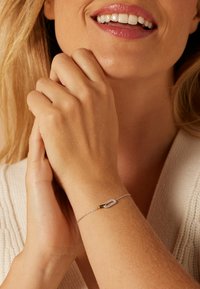 Zilveren armband met een decoratieve heldere stenen accent in rechthoekige vorm, voorzien van een goudkleurige sluiting. Subtiele textuur op de huid.