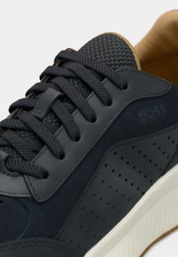 Lähivõte tumelainest spordijalatsist, millel on tekstuuriga paelad, perforatsiooniga külgpaneelid ja ülaosas nähtav BOSS-logo.