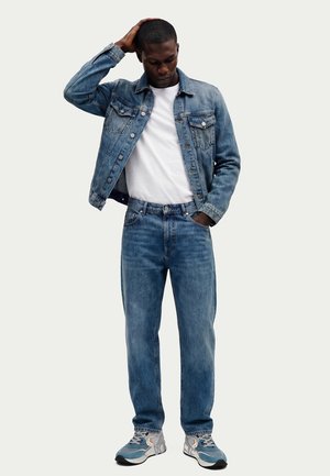 Uomo con giacca di jeans blu e jeans, t-shirt bianca e sneakers, in piedi con una mano sulla testa e l'altra nella tasca.