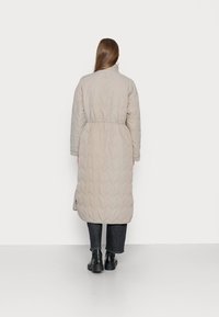 Abrigo largo acolchado en color beige con cintura ajustable, cuello alto y patrón texturizado en chevron. Lleva puesto un par de jeans oscuros y botas negras.