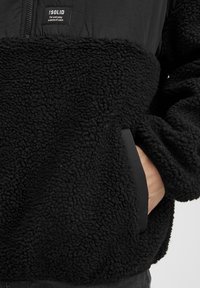 Svart fleece-pullover med en texturerad, fluffig tyg, med dragkedja framtill och sidofickor, kompletterad med en slät svart panel vid fållen.