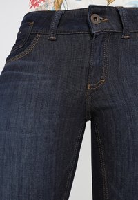 Dark blue denim jeans featuring a fitted design, visible front pocket details, and contrasting stitching along seams and button closure.