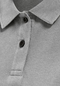 Lysegrå strikket poloshirtkrave med to sorte knapper på en tekstureret stofbaggrund.