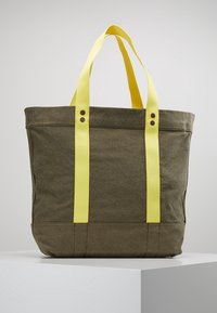 Olivgrön kanvas tote-väska med gula remmar, stor rektangulär form, förstärkt botten och metallknappar på sidorna.