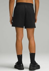 Shorts de sport noirs avec une taille plate, un tissu lisse et une coupe décontractée ; portés avec des chaussettes noires et des chaussures de course.