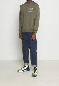 Olivově zelená mikina s textem, žebrovanými manžetami a malým bílým logem Nike. Spárována s ohrnutými tmavě modrými džíny a vícebarevnými teniskami.