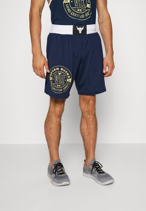 Granatowe szorty sportowe z nadrukiem logo w kolorze żółtym, elastycznym pasem i kontrastującym białym paskiem. Połączone z szaro-pomarańczowymi sneakersami.