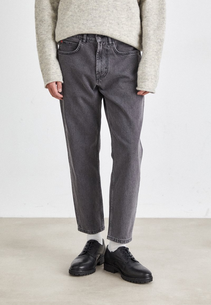 Jean en denim gris avec une coupe droite, taille moyenne et design classique à cinq poches, associés à des chaussures à lacets en cuir noir.