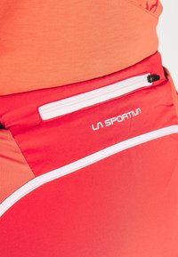 Červené sportovní šortky s kapsou na zip a bílými akcenty; síťovinová textura po stranách; značení viditelné na pásku.