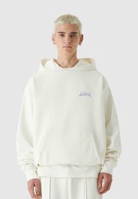 Sudadera blanca con capucha y bolsillo delantero, de textura suave y con un logo morado claro en el pecho. Corte relajado y mangas largas.