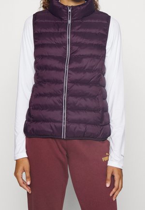 Osoba ubrana w ciemnofioletową pikowaną kamizelkę na białej koszuli z długim rękawem i bordowe spodnie dresowe Puma, stojąca z rozluźnionymi ramionami.