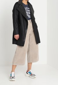 Černý kabát z umělé kůže s texturovaným šedým límcem, doplněný béžovými širokými kordovými kalhotami a vícebarevnými sportovními teniskami.