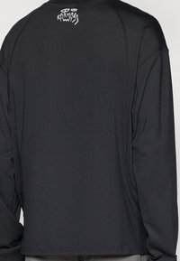 Black long-sleeve shirt with a relaxed fit, featuring a white graphic logo on the back and flatlock seams for a smooth finish.