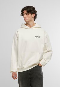 Felpa color crema realizzata in tessuto morbido, con tasca a canguro e testo nero sul lato sinistro del petto. Design comodo con maniche ampie.