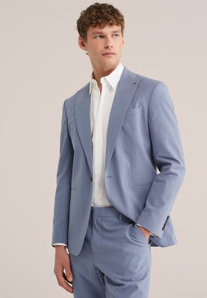 Mladý muž v svetlomodrom obleku a bielej košeli, stojaci s jednou rukou v vrecku, pozerajúci sa do strany na neutrálnom pozadí.