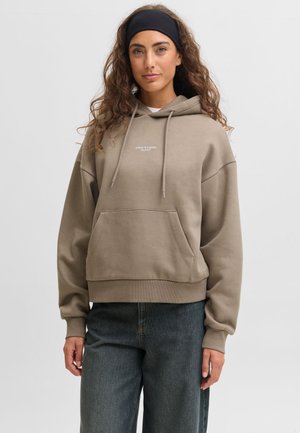 Femme aux cheveux bouclés portant un bandeau noir, un sweat à capuche beige avec poche avant et un jean bleu foncé, debout devant un fond gris uni.