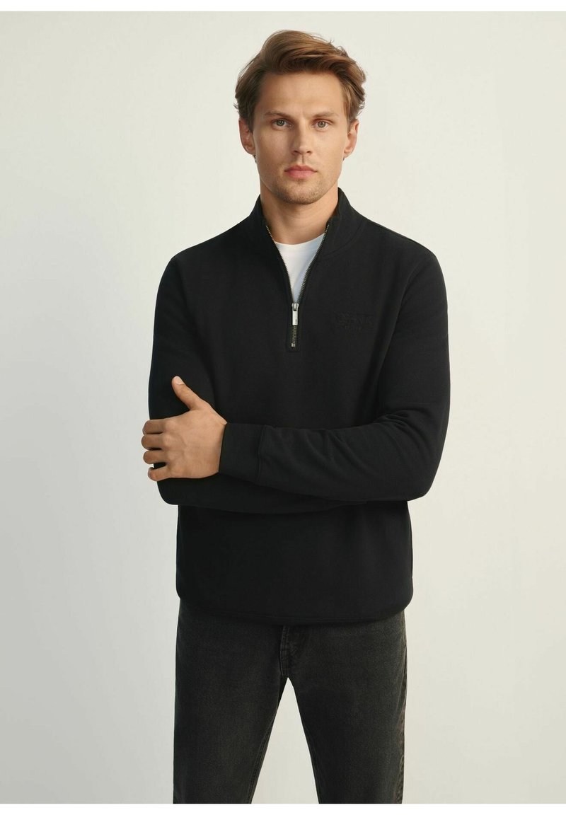 Czarny pulower z ćwierć zamkiem, wykonany z gładkiego materiału, z małym logo na piersi, długimi rękawami i luźnym krojem.