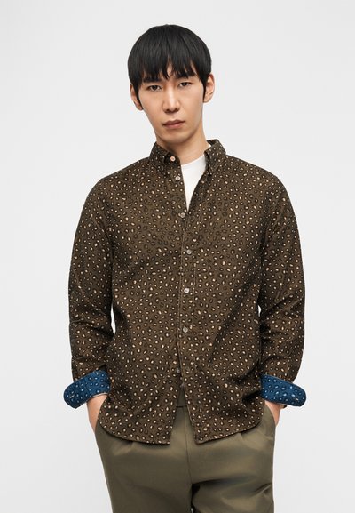 Άνδρας που φοράει καφέ πουκάμισο με κουμπιά και τύπωμα λεοπάρδαλης, με μπλε μανσέτες, λευκό μπλουζάκι και ελαιί πράσινο παντελόνι, στέκεται με τα χέρια στις τσέπες.