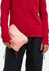 Rode, gestructureerde fleecetrui met een volledige rits en aansluitende manchetten; houdt een roze, opvouwbare donsjack met logo vast. Zwarte broek op de achtergrond.