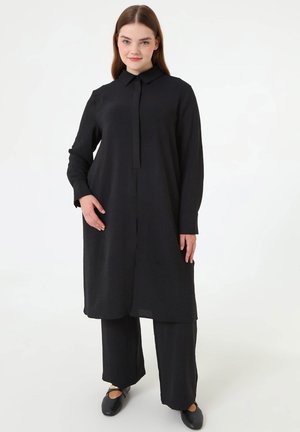 Jonge vrouw met stijl bruin haar, gekleed in een lange zwarte hemdjurk en brede zwarte broek, staand tegen een witte achtergrond.