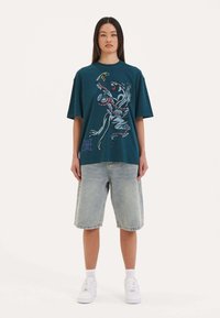 Teal oversized t-shirt med et farverigt grafisk design, parret med lyse denimshorts og hvide sneakers. Casual, afslappet fit.