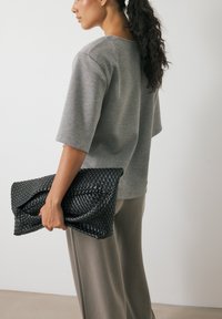 Juodas austas odinis kosmetinis krepšys laikomas vienoje rankoje, derinamas su pilku trumpu rankovėmis marškinėliu ir bežinėmis plačiomis kelnių. Lygūs paviršiai ir struktūrizuotas dizainas.