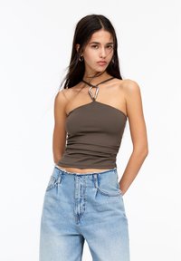 Hnedý priliehavý crop top s jedným ramienkom a strieborným kruhovým akcentom. Skombinovaný s vysoko pasovými svetlomodrými džínsami.