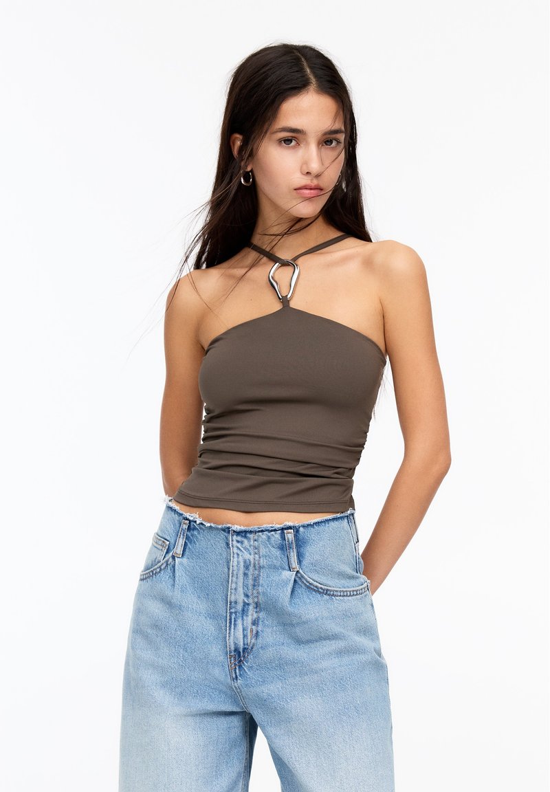 Hnedý priliehavý crop top s jedným ramienkom a strieborným kruhovým akcentom. Skombinovaný s vysoko pasovými svetlomodrými džínsami.