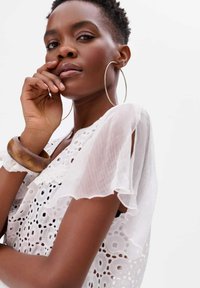 Biała koronkowa bluzka z falbaniastymi rękawami, ozdobiona kwiatowymi wzorami i wycięciami. Duże metalowe kolczyki w kształcie kół oraz drewniana bransoletka.