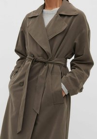 Persoon draagt een olijfgroene trenchcoat met riem over een lichtgrijze trui, met handen in de jaszakken tegen een effen achtergrond.