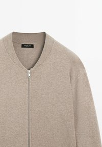 Cardigan beige con zip realizzato in tessuto morbido. Presenta un colletto e polsini a coste, con una texture liscia e dettagli di cucitura minimi.