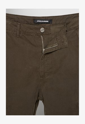 Pantaloni maro cu fermoar metalic parțial desfăcut, nasture cu marcă și etichetă „Pegador” pe talie.