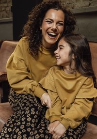 Bijpassende mosterdgele sweatshirts met rimpeldetails; vrouw en meisje zittend op een bruine bank, met zwart-gepatroonneerde broek met bloemenpatronen.