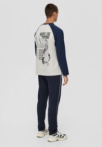 Langärmliges Shirt mit marinefarbenen Ärmeln und einem hellgrauen Körper, ausgestattet mit einem Rückendruck mit Text und Design. Kombiniert mit marinefarbenen Hosen und Sneakers.