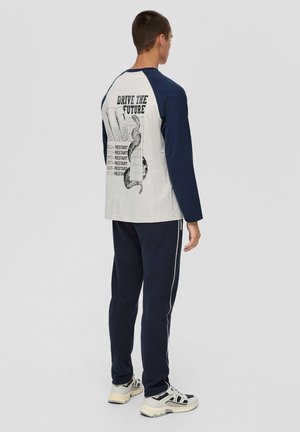 Langsleeve shirt met marineblauwe mouwen en een lichtgrijze romp, met een achterafdruk met tekst en een ontwerp. Gecombineerd met marineblauwe broek en sneakers.
