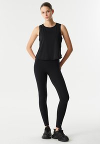 Zwart mouwloos topje met een losse pasvorm, gecombineerd met zwarte leggings. Schoeisel bestaat uit zwarte sportschoenen. Eenvoudige, gladde stof zonder patronen.