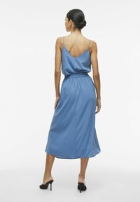 Robe midi en satin bleu avec décolleté en V, fines bretelles, taille élastique et jupe fluide. Portée avec des chaussures à talons hauts noires.