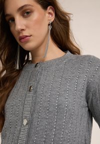 Szary kardigan z pionowym prążkowaniem, ozdobiony małymi srebrnymi ćwiekami i dwoma guzikami. Długie, srebrne kolczyki z frędzlami dopełniają stylizację.