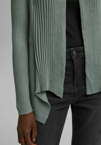 Vihreä ribbineulottu neuletakki, jossa leveä avoin etuosa ja epäsymmetrinen helma. Pehmeä pinta, pitkät hihat ja näkyvät sivutaskut. Mustat housut alla.