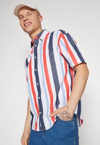 Camicia con bottoni caratterizzata da strisce verticali rosse, blu navy e azzurro chiaro, maniche corte e un piccolo logo. Materiale in cotone con una vestibilità casual.