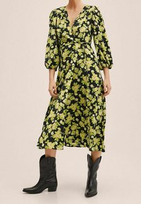 Robe noire avec de courtes manches bouffantes, ornée d'un imprimé floral jaune-vert. Longueur genou, design portefeuille, associée à des bottes de cowboy noires.