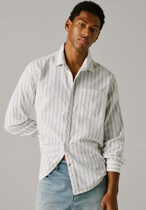 Noor mees, kes kannab valget pikavarrukalist särk õhukeste vertikaalsete triipudega ja helesiniseid teksapükse, seisab ühevärvilise halli tausta ees.