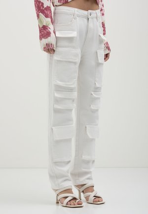 Weiße Cargo-Hosen mit mehreren Klappentaschen, kombiniert mit weißen High-Heel-Sandalen und einem kurz geschnittenen, blumigen Sweater.