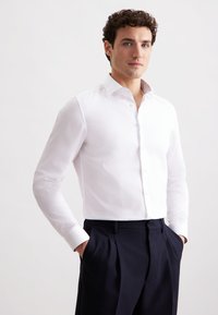 Weißes Langarmhemd mit spitzem Kragen, das einen gemusterten Innenkragen aufweist, kombiniert mit maßgeschneiderten navy-farbenen Hosen. Sanfte Textur.