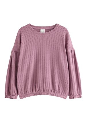 Ung pige i mauve strikket outfit og hue, bærende en lilla rygsæk med snøre og tegneserie dyreprint, smilende og kiggede sidelæns.