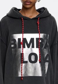 Felpa con cappuccio in cotone grigio scuro con una grafica metallica argentata che riporta la scritta "BIMBA Y LOLA". Presenta laccetti con motivo rosso e bianco.